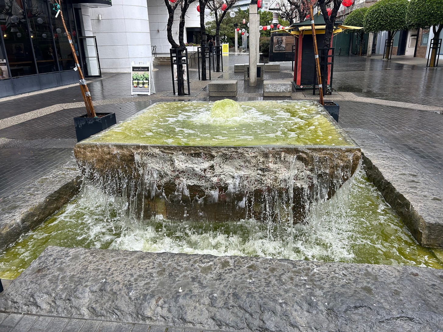 A ground fountain, square-shaped, emanating green foamy liquid in a concrete plaza