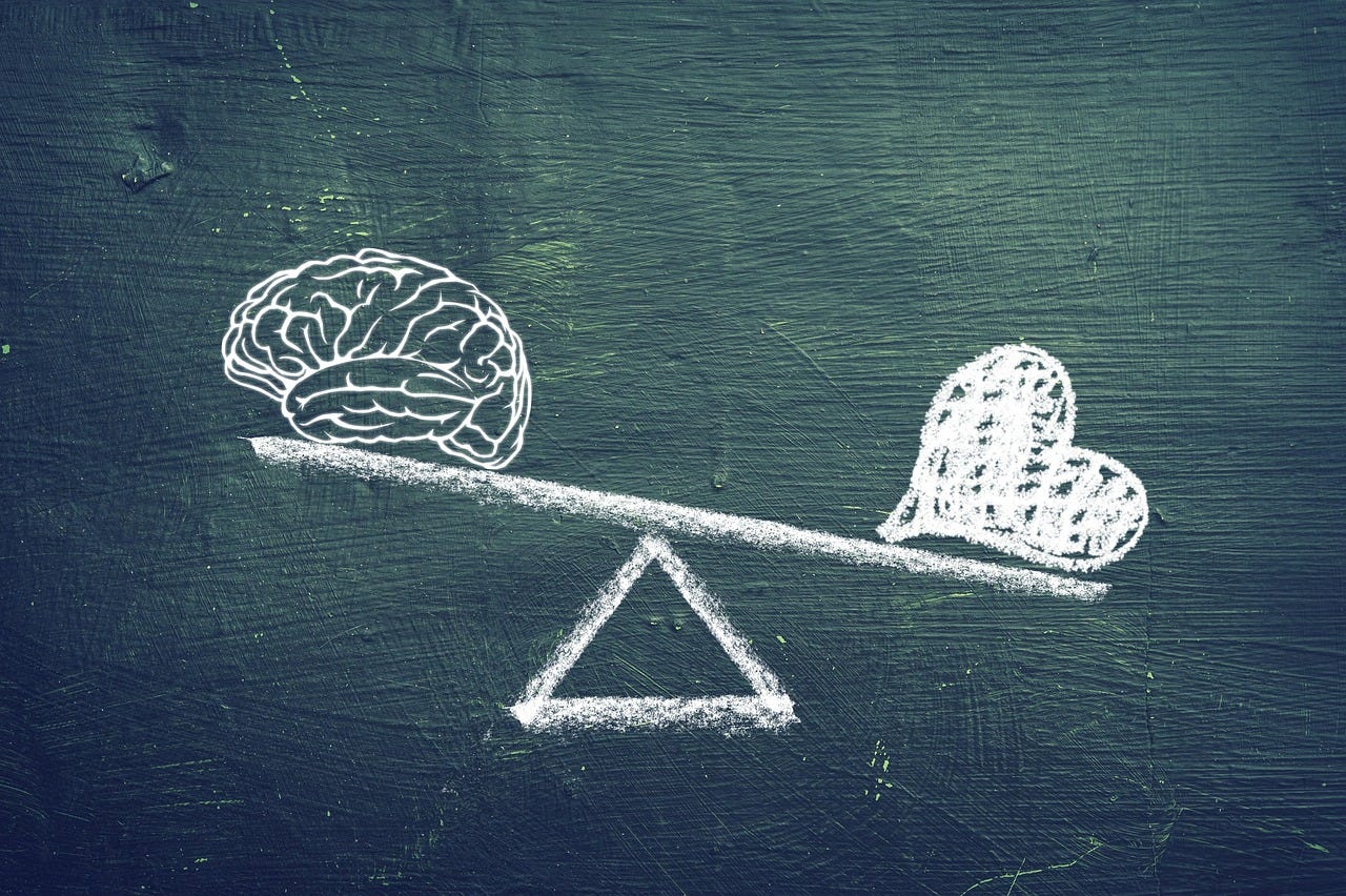 Balance scale with a brain on the left side, raised, and a heart shape on the right side, lowered. Drawn in white chalk on a dark board.