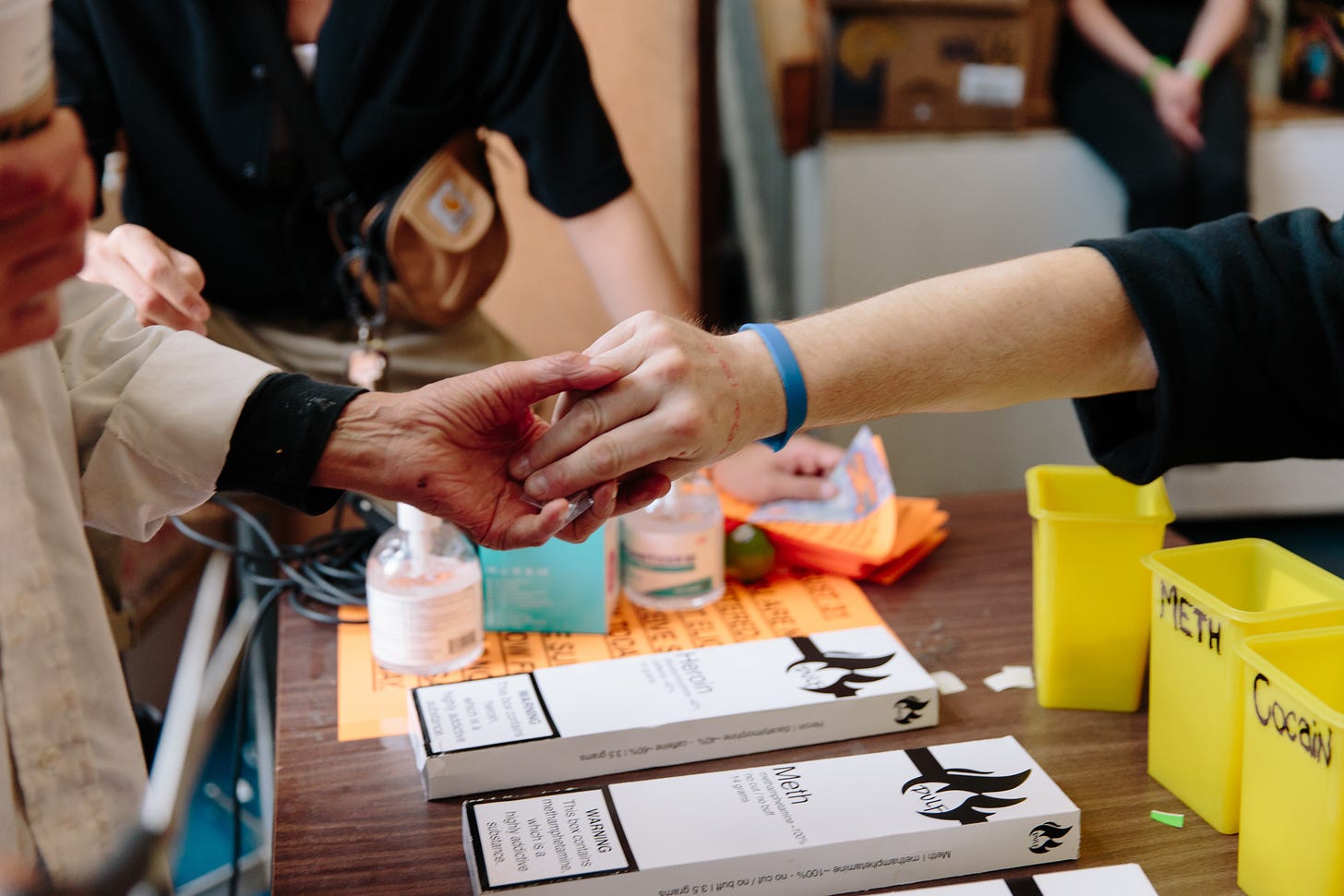 hands exchange what appears to be a baggie containing drugs over a wooden desk, on which sits plastic yellow buckets labelled "Meth" and "Cocaine". Regulated labels that read "Heroin" and "Meth" sit on the table as well, showing the purity of the tested substances, beside two bottles of hand sanitizer. The hands slightly grasp one another, in an affectionate way indicating familiarity.