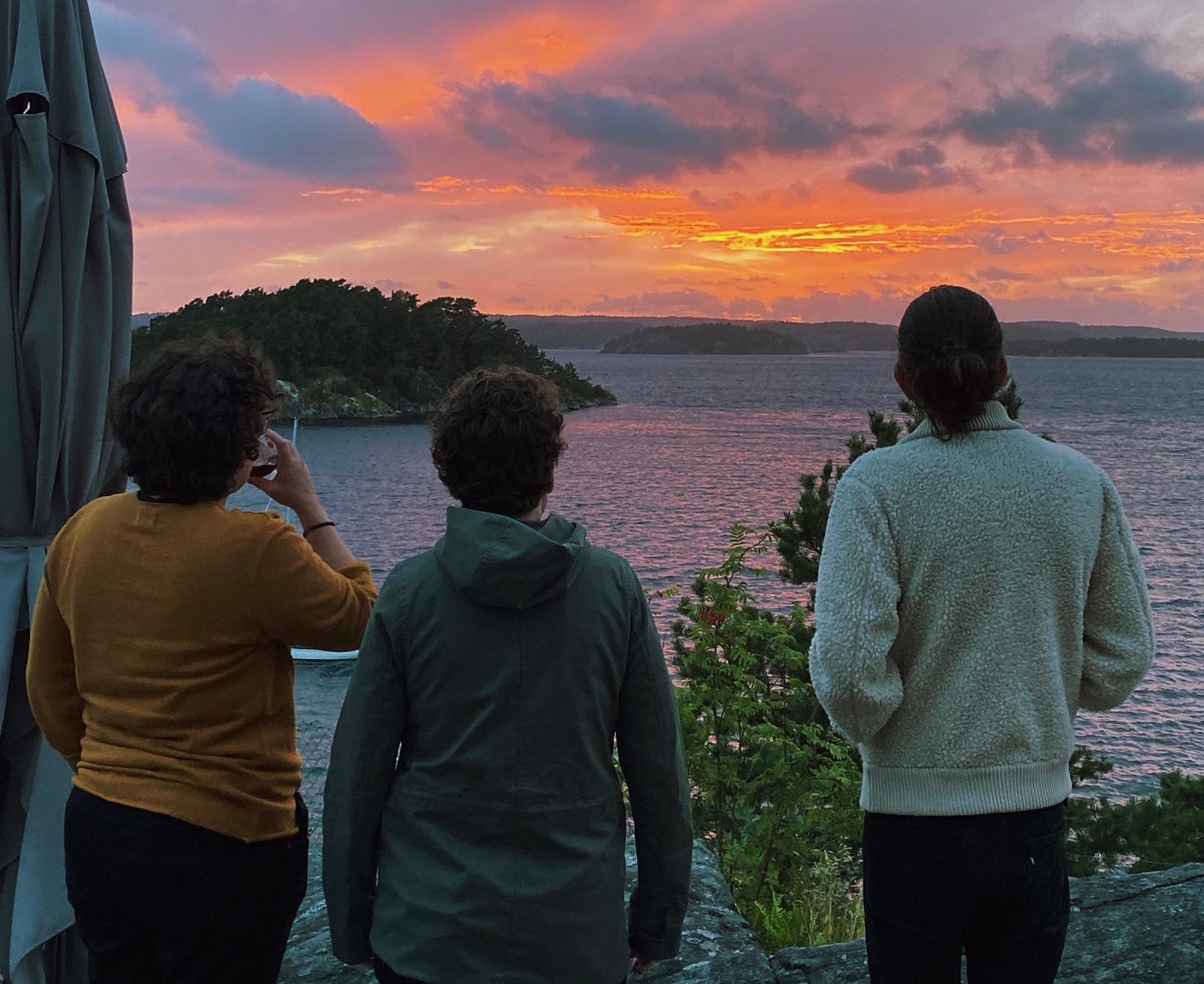 Três pessoas de costa olhando para o pôr do sol. À frente o mar, uma ilha e o pôr do sol bem alaranjado e com bastante nuvens formando uma paisagem dramática.