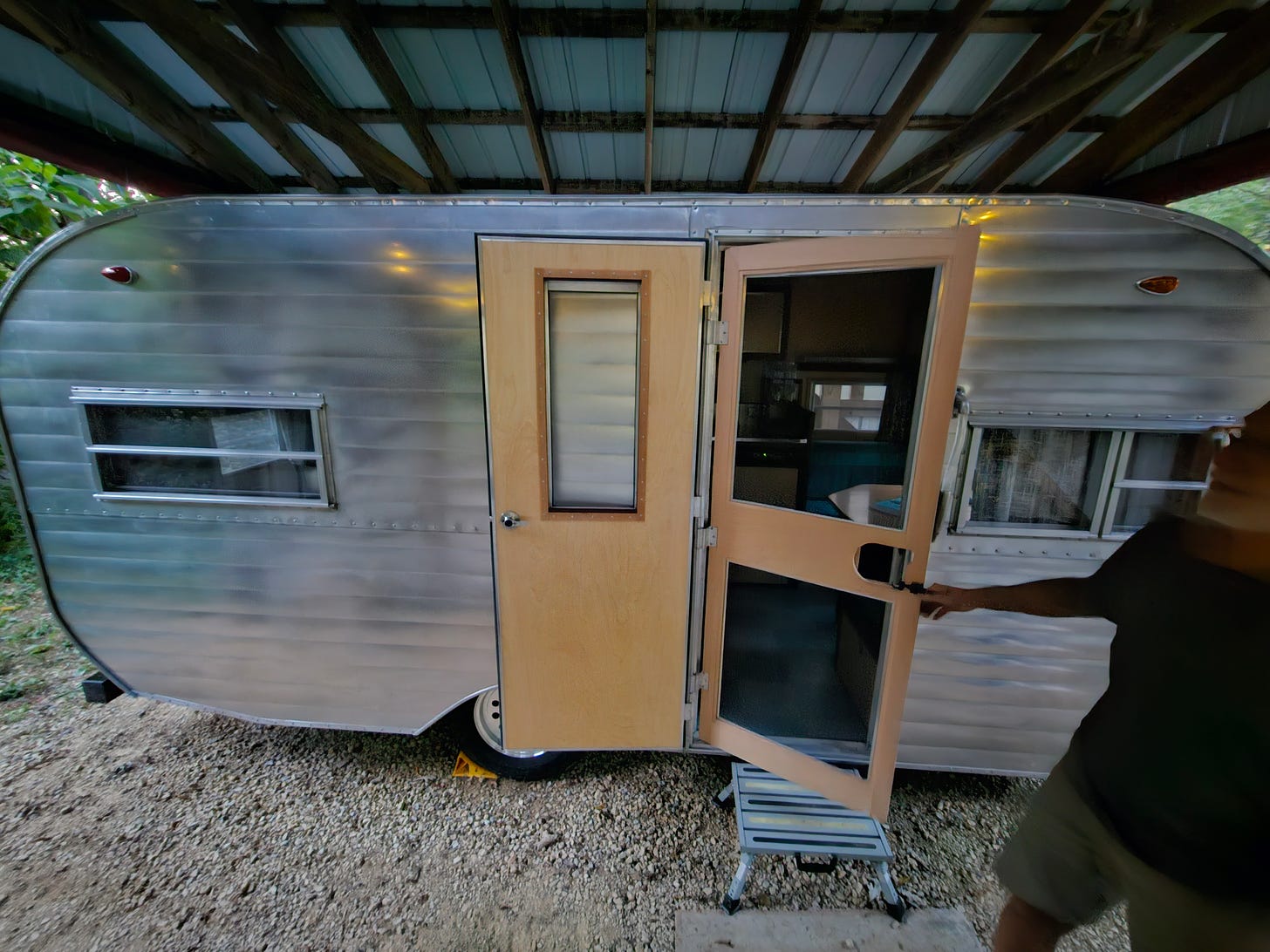 A silver metal, canned-ham-style camper trailer has a lovely wooden screen door that's being opened by an older white man with a silver beard.