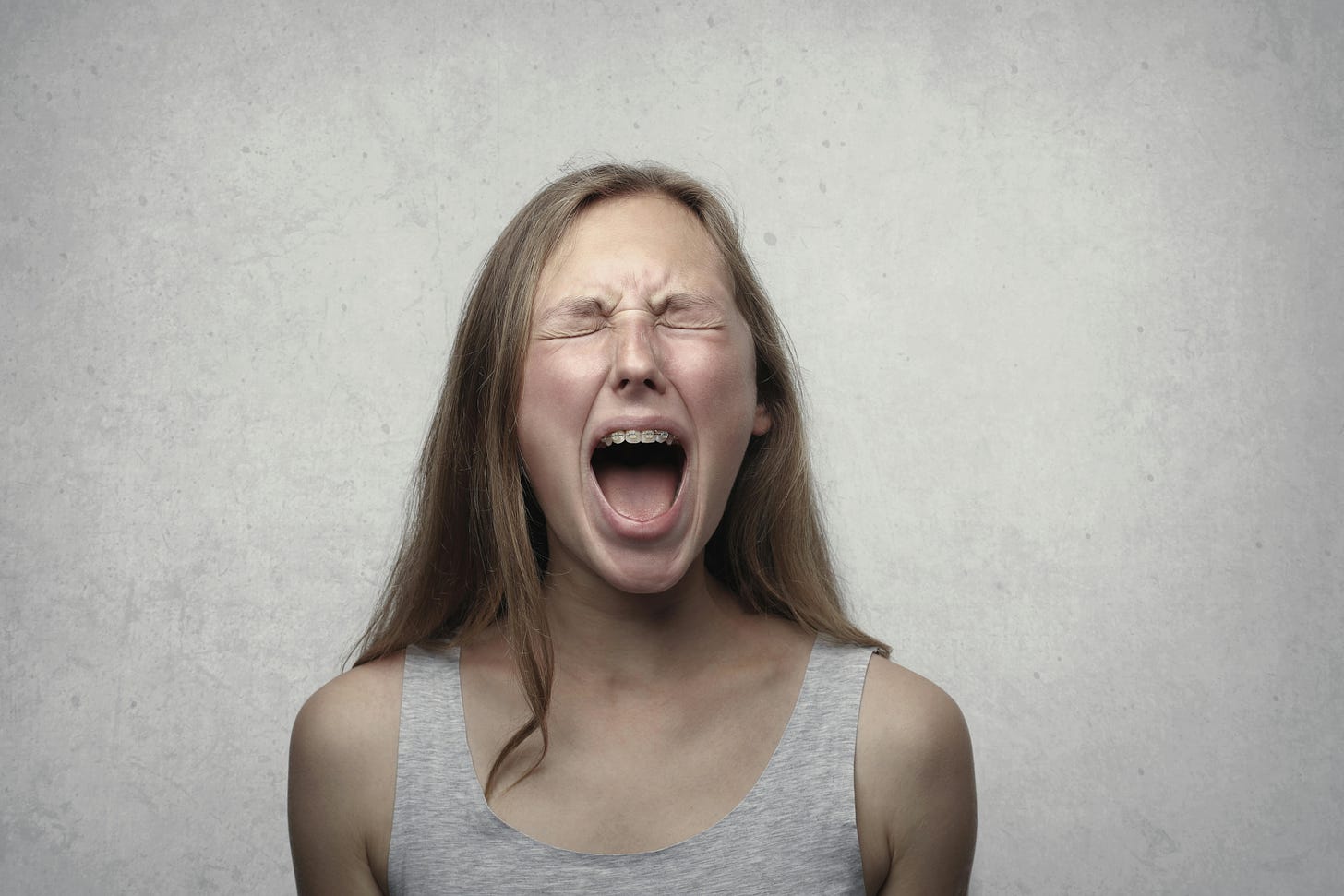 Photo by Andrea Piacquadio: white woman with dark blonde hair and in grey tank top opening her mouth and shutting her eyes like she is screaming out in pain. There is a grey background in this photo and she is wearing braces.