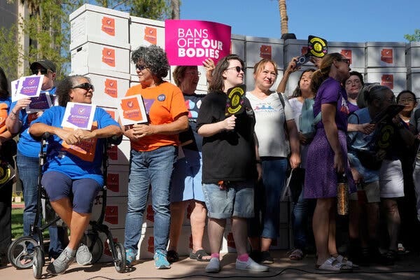 A group of people stand together and hold signs. One reads “Bans Off Our Bodies.” Stacked behind them are white boxes with stickers that read “Arizona for Abortion Access.”