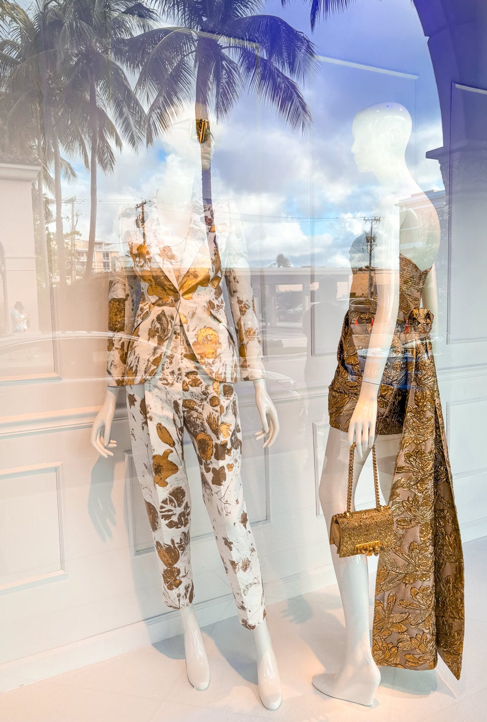 Shop window with two mannequins wearing white and gold outfits with palm trees reflected in glass