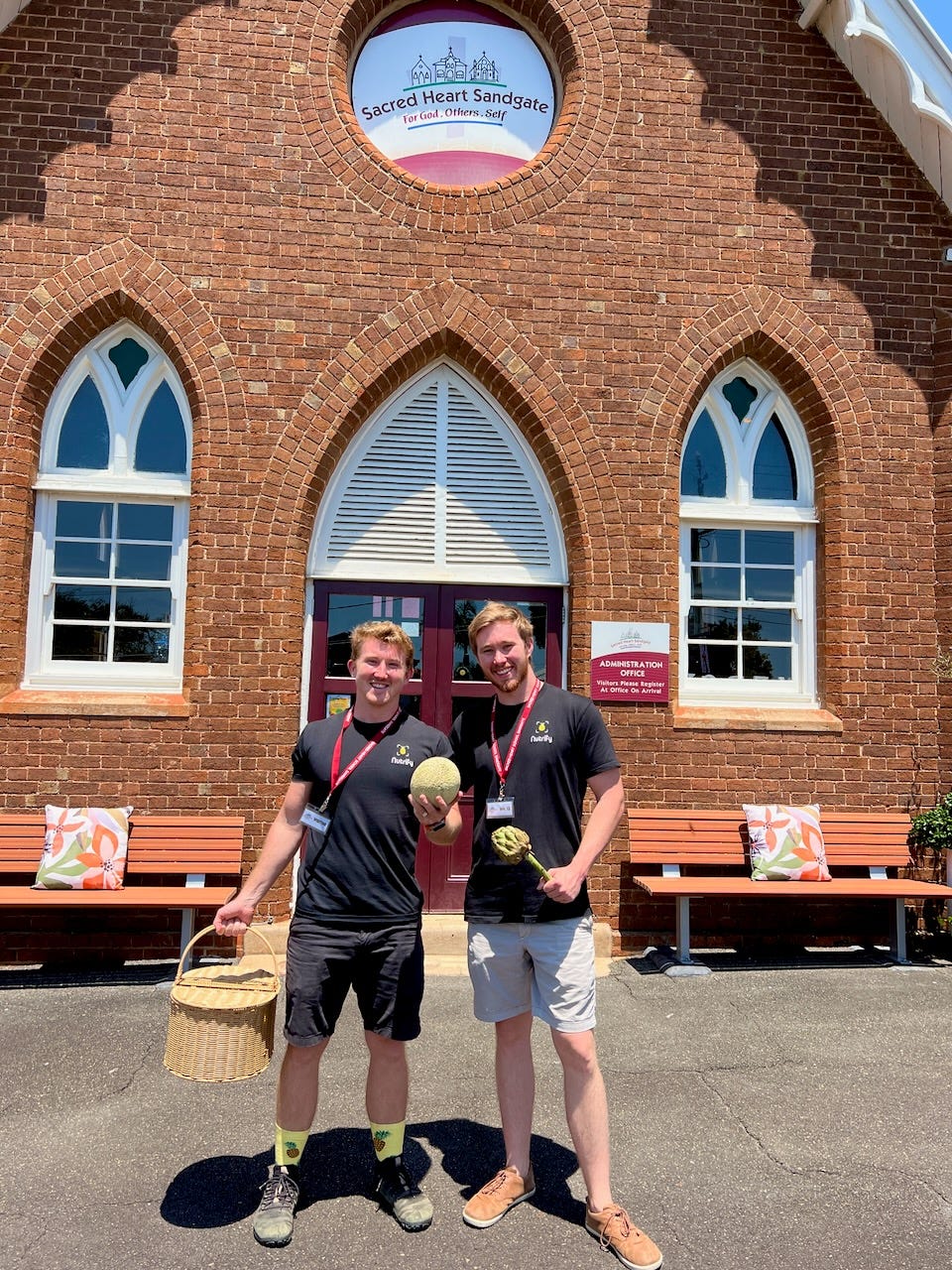 Two men wearing black Nutrify shirts and red lanyards stand smiling in front of Sacred Heart Sandgate, a brick building with arched windows and a sign above the entrance. One holds a basket and the other holds a melon and artichoke. They stand on a paved surface, with benches and decorative cushions on either side of the entrance.