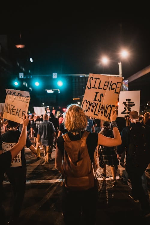 Picture from the back of a protest, showing a girl in a black t-shirt and tan backpack holding up a sign written on a piece of cardboard that says "Silence is Compliance".
