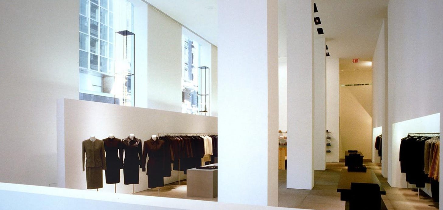 A grainy film photograph depicts an airy space with white walls and columns with racks of minimalist clothes to the left and windows with light streaming in above. 