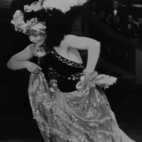 Animated gif - silent star Gilda Gray dances the shimmy in 1929 film Piccadilly. She’s wearing an elaborate cabaret costume with a feathered headdress and a swishy skirt