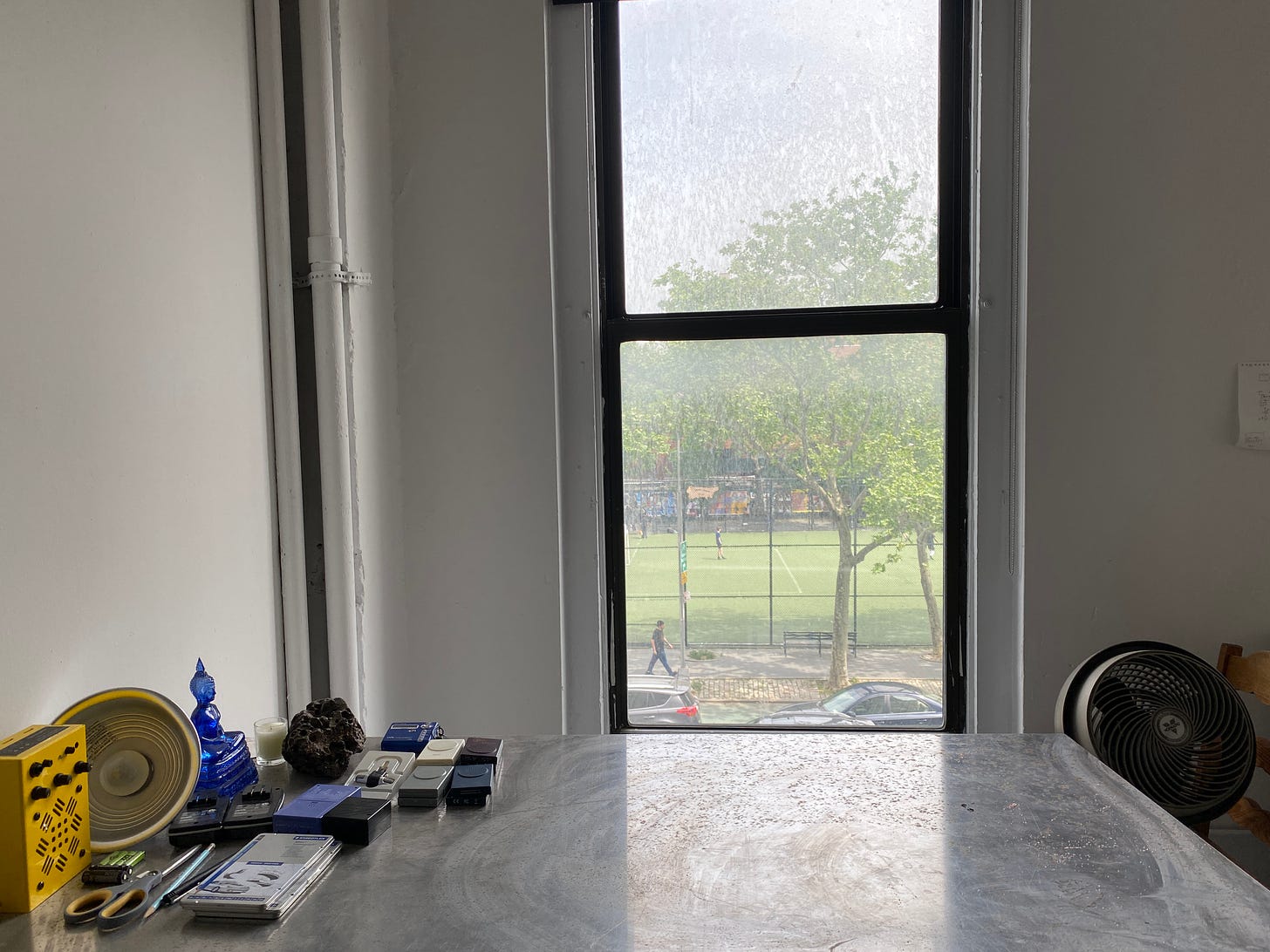 A stainless steel work table in front of a window. On the table to the left are a box of pencils, a pair of scissors, a yellow speaker-amplifier, an aluminum clip light, a stone with holes in it, and a translucent blue resin Buddha. Out the window are green soccer fields and leafy trees. You can see a few cars and a person walking.