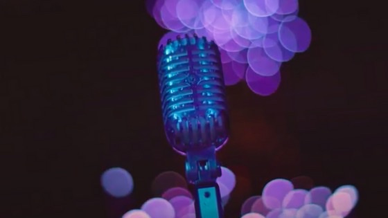 A high tech microphone reflects the turquoise and electric blue lights. In the background, lavender globes glow in the black.