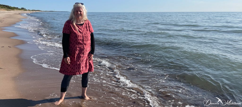 Daniela Mühlheim, the author of this substack, stands barefoot in the Baltic Sea smiling in a ruby coloured dress