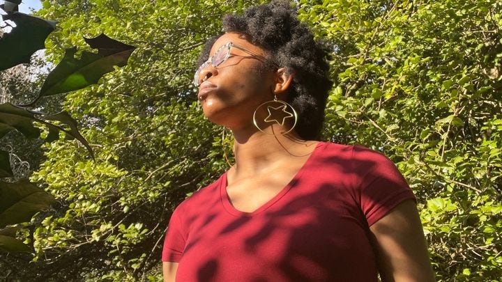 A Black trans disabled artist is closing their eyes and taking in the sun, against the green leaves of a tree. They are wearing gold hoop earring with a star in the middle and a red t-shirt.