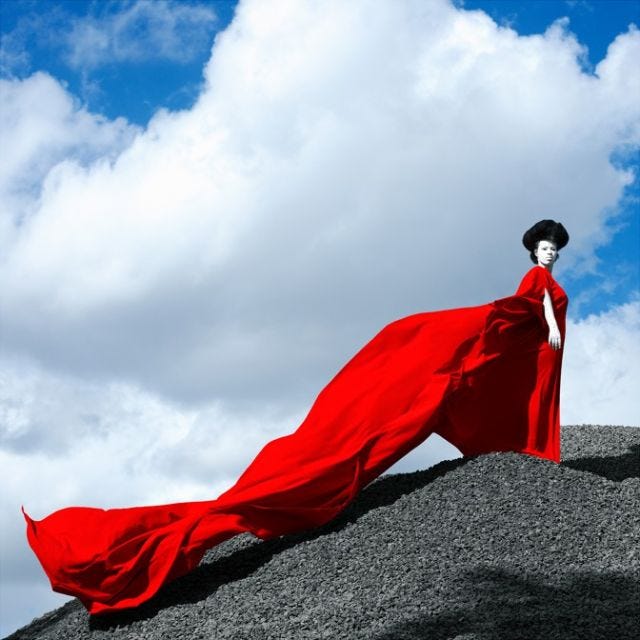 A woman with white painted skin and black hair wears a flowing red dress while standing on top of a grey rock