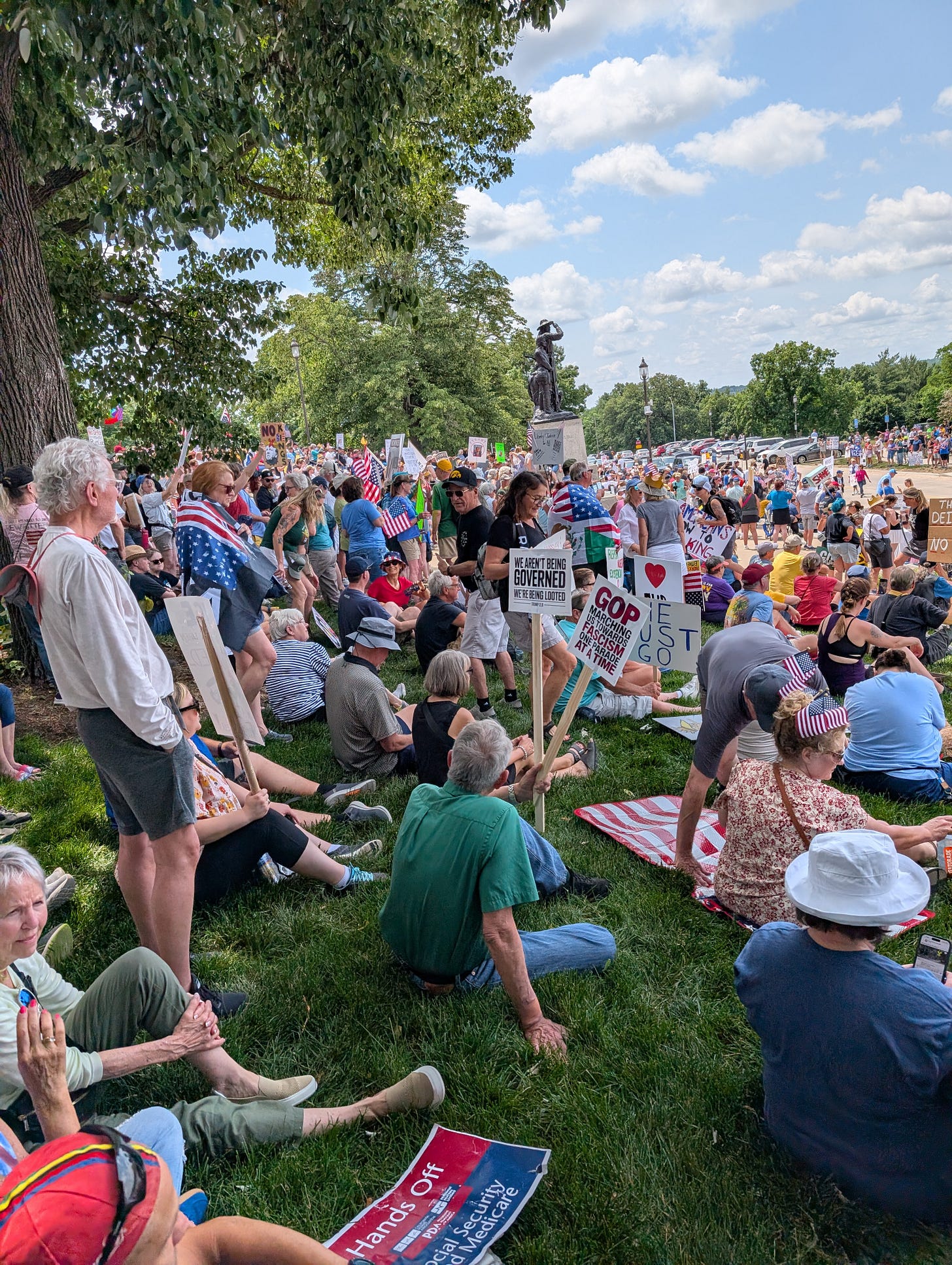 A crowd, featuring multiple AMerican flags and holding signs that say things like "We aren't being governed, we're being looted" and "GOP marching us towards fascism" gathers under trees in a park around a monument. The park is packed with people as far as the eye can see. A crowd, featuring multiple AMerican flags and holding signs that say things like "We aren't being governed, we're being looted" and "GOP marching us towards fascism" gathers under trees in a park around a monument. The park is packed with people as far as the eye can see.