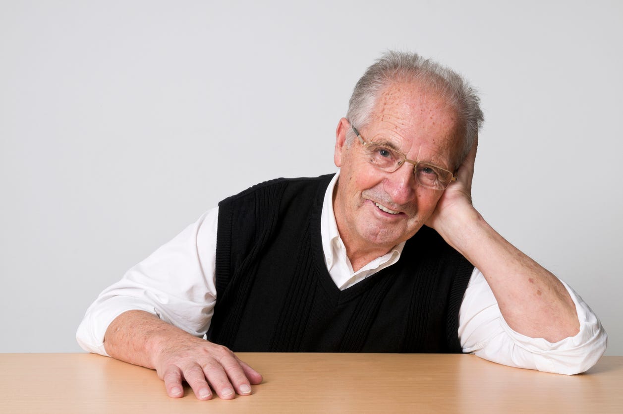 Smiling, balding white man rests  his left elbow on table in front of him and has his palm along side of his face. He's wearing a white shirt and black vest.  