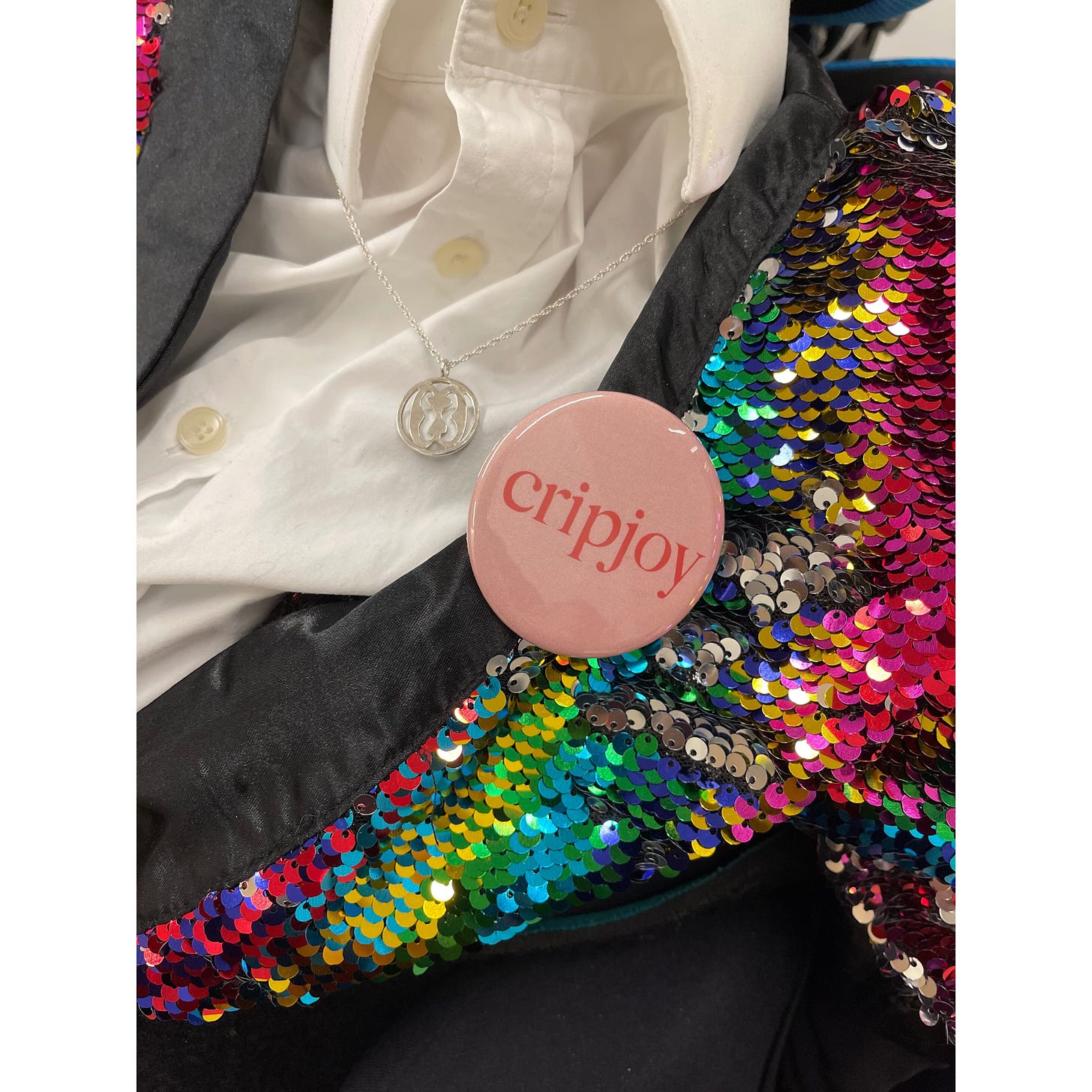 IMAGE DESCRIPTION 7: A close up of Sulaiman’s necklace and badge against his white dress shirt and rainbow coloured sequin kimono.