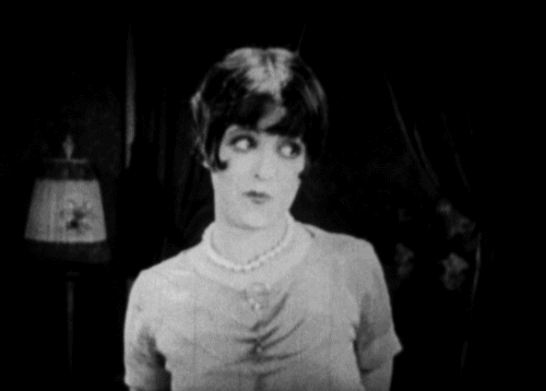 A black and white image of Clara Bow. She's looking off to her right smiling and raises her hands to her head like a moose and wiggles her fingers in a ner her way.