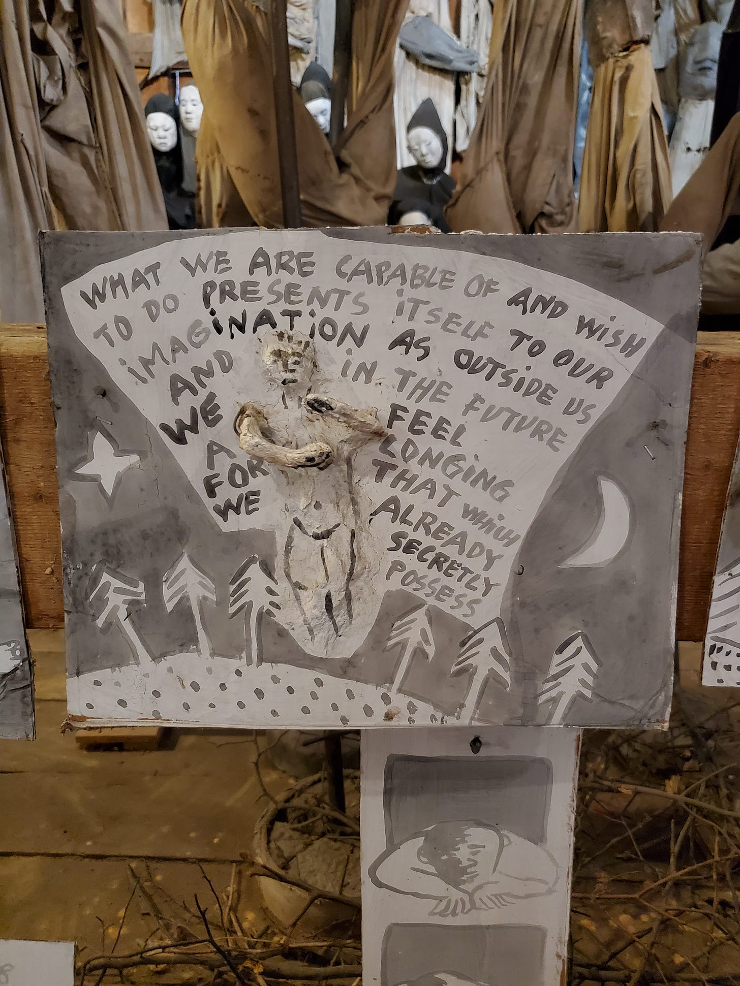 A grayscale hand-painted sign with a three-dimensional figure in the center reads "What we are capable of and wish to do presents itself to our imagination as outside us and in the future we feel a longing for that which we already secretly possess"