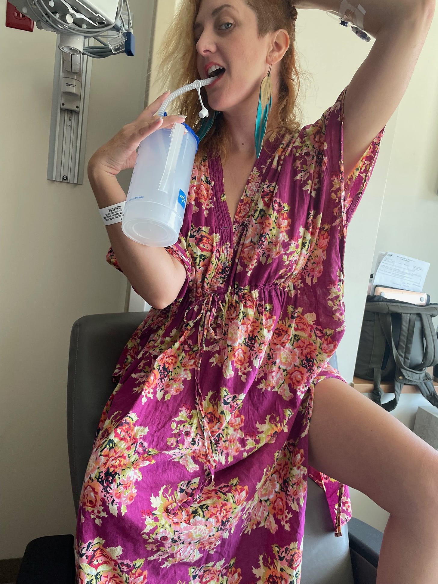 A white woman with red hair wearing a floral dressing gown poses suggestively with a hospital-issued water bottle in a hospital room.