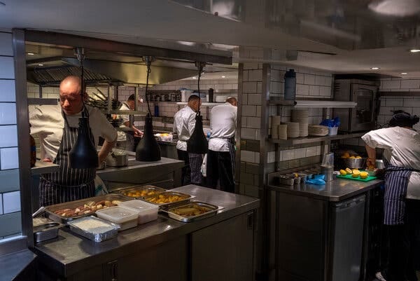 A view of the kitchen, with food laid out in stainless steel trays and chefs in aprons at work. In one corner a woman is cutting lemons.