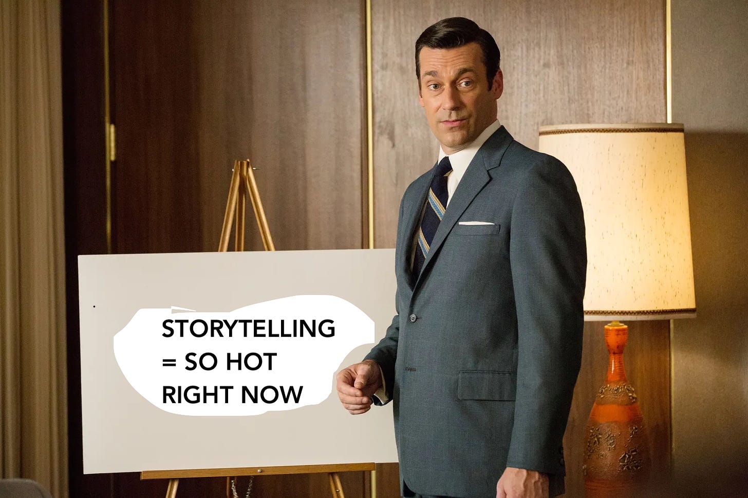 A 1960's ad executive named Don Draper, in a gray suit, looks a little confused in a wood paneled room. He's standing in front of an easel. I've edited the easel to say: "Storytelling = So Hot Right Now."