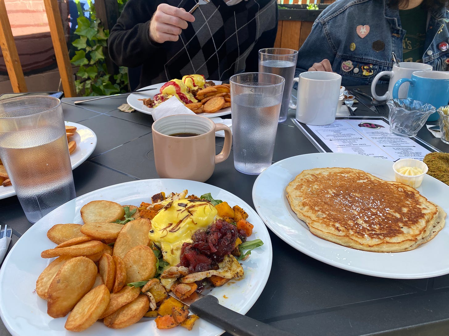 A patio table with plates of breakfast foods and cups of coffee and water. In the foreground is a plate with fried slices of potato, a half benny with yams and chutney and arugula, and another plate with a large pancake and a side of whipped butter. My brother Riley is visible in the background, his hand holding a fork over his plate.