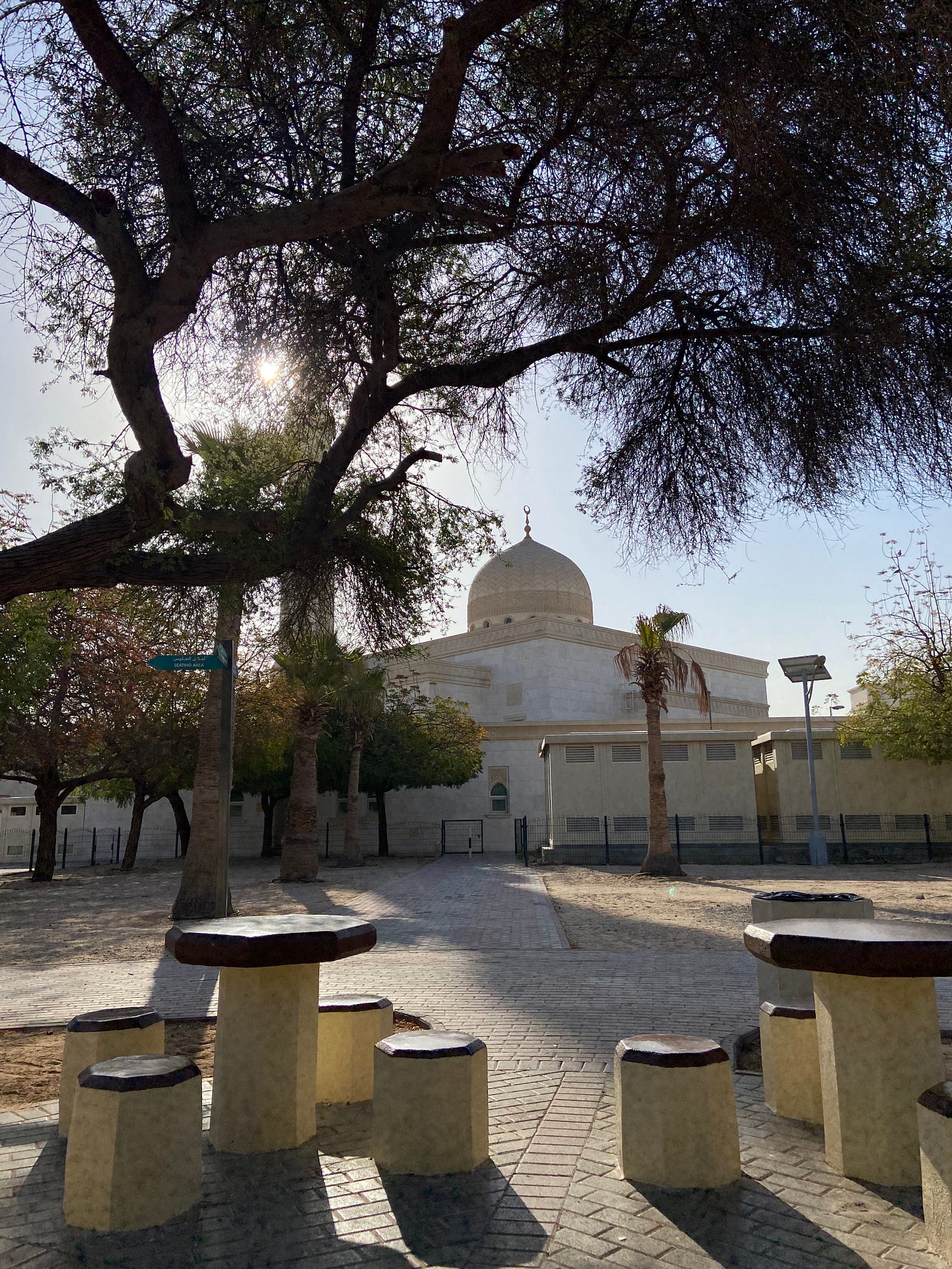 Photo of a small park during mid-morning with hexagonal shaped table and hexagonal shaped thick stools and an ivory colored mosque in the background