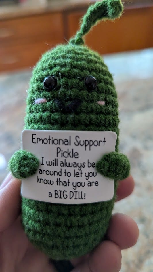 Crocheted green pickle with cute face holding a sign, "emotional support pickle, I will always be around to let you know that you are a big dill!"