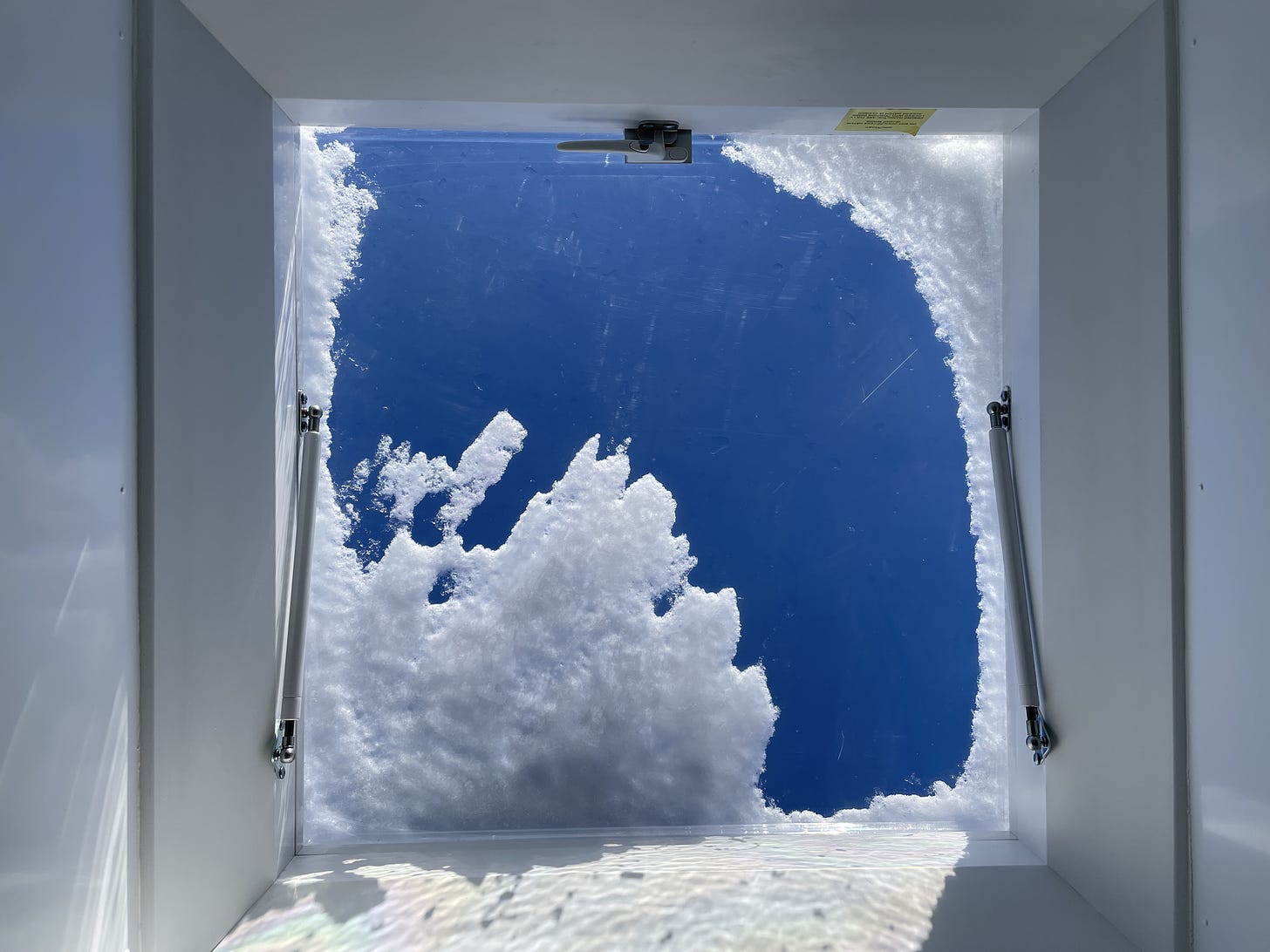 Looking up at a roof access hatch. There are blue skies and snow is resting on the plastic hatch.