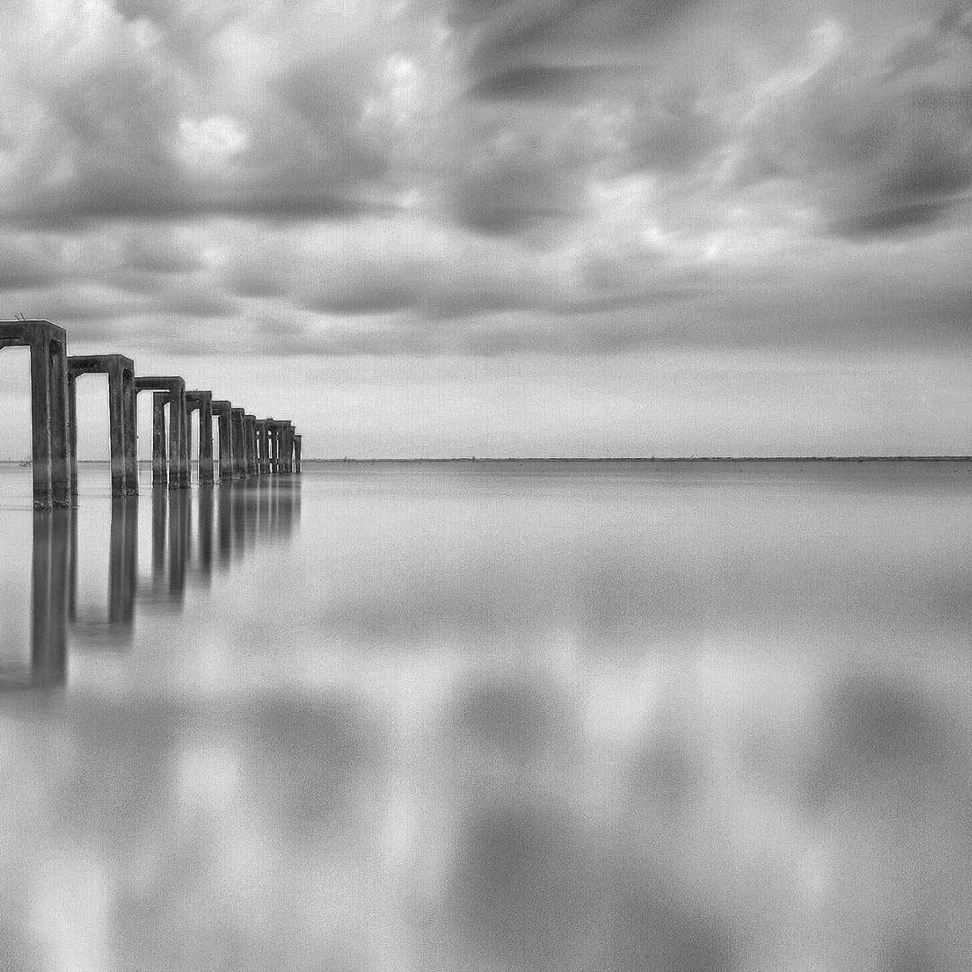 A photo of a grey water reflecting a grey cloudy sky, with a series of archlike structures to one side