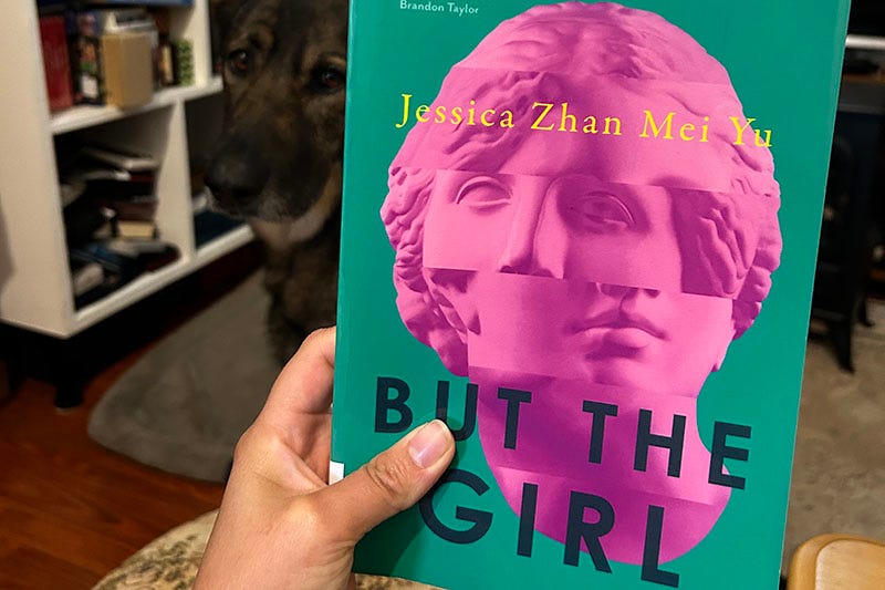 A hand holds a copy of 'But The Girl' by Jessica Zhan Mei Yu, while a dog looks on.