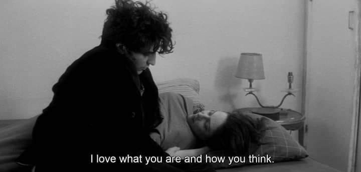 This may contain: a woman laying on top of a bed next to a man in a black jacket