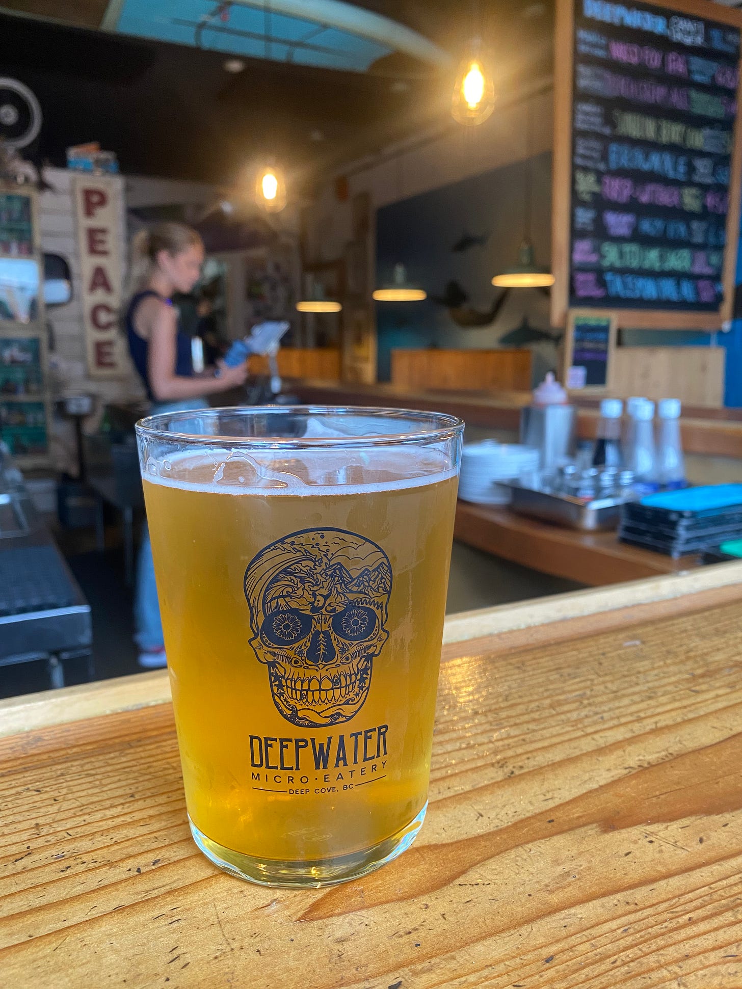 A glass of beer with a skull logo on it, on a wooden bartop. Blurred in the background is the rest of the small bar, a bartender standing at a register, a beer list on a chalkboard, some decorative signs, a couple of tables.