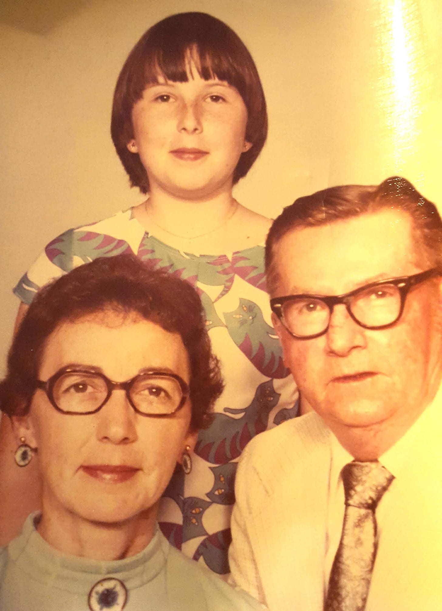 Seventies era family photo. Seated at left: dark haired woman with dark glasses, smiling slightly. To her left, gray haired man with dark glasses, attentive expression. Standing over them on a platform: smiling girl wearing a dress with a purple and blue tabby cat motif.