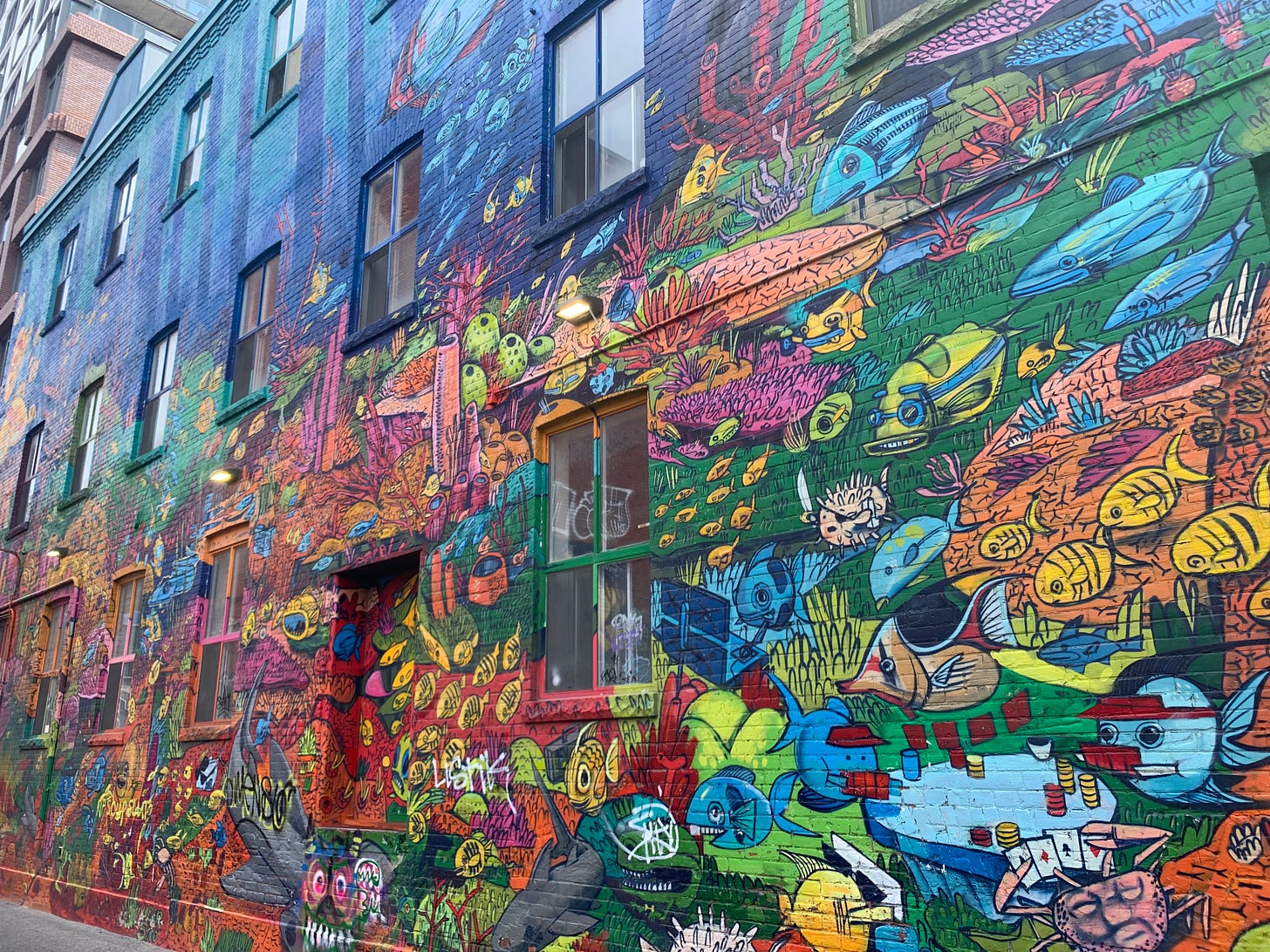 A colourful mural with a variety of cartoon fish and coral in bright yellow, orange, green, and pink, some swimming and others doing various human things. The top of the image transitions into a variety of blue hues.