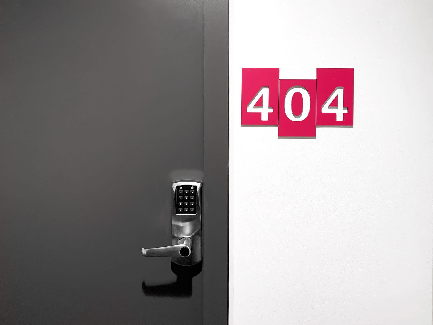 uma porta cinza, com a maçaneta com um teclado numero. Ao lado, numa parede branca, escrito com fundo vermelho e letras brancas está o número do quarto: 404.