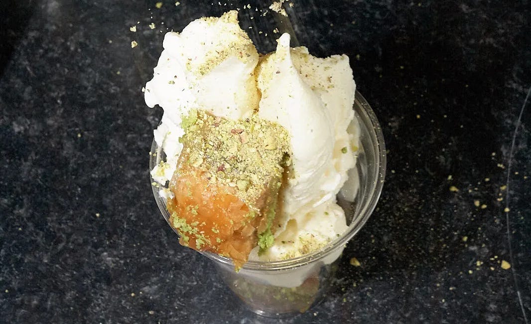 On a black granite countertop is a clear plastic cup filled with two peaks of luscious white ice cream, with a baklava nestled on the side, while pistachio nut crumbles are sprinkled from above.