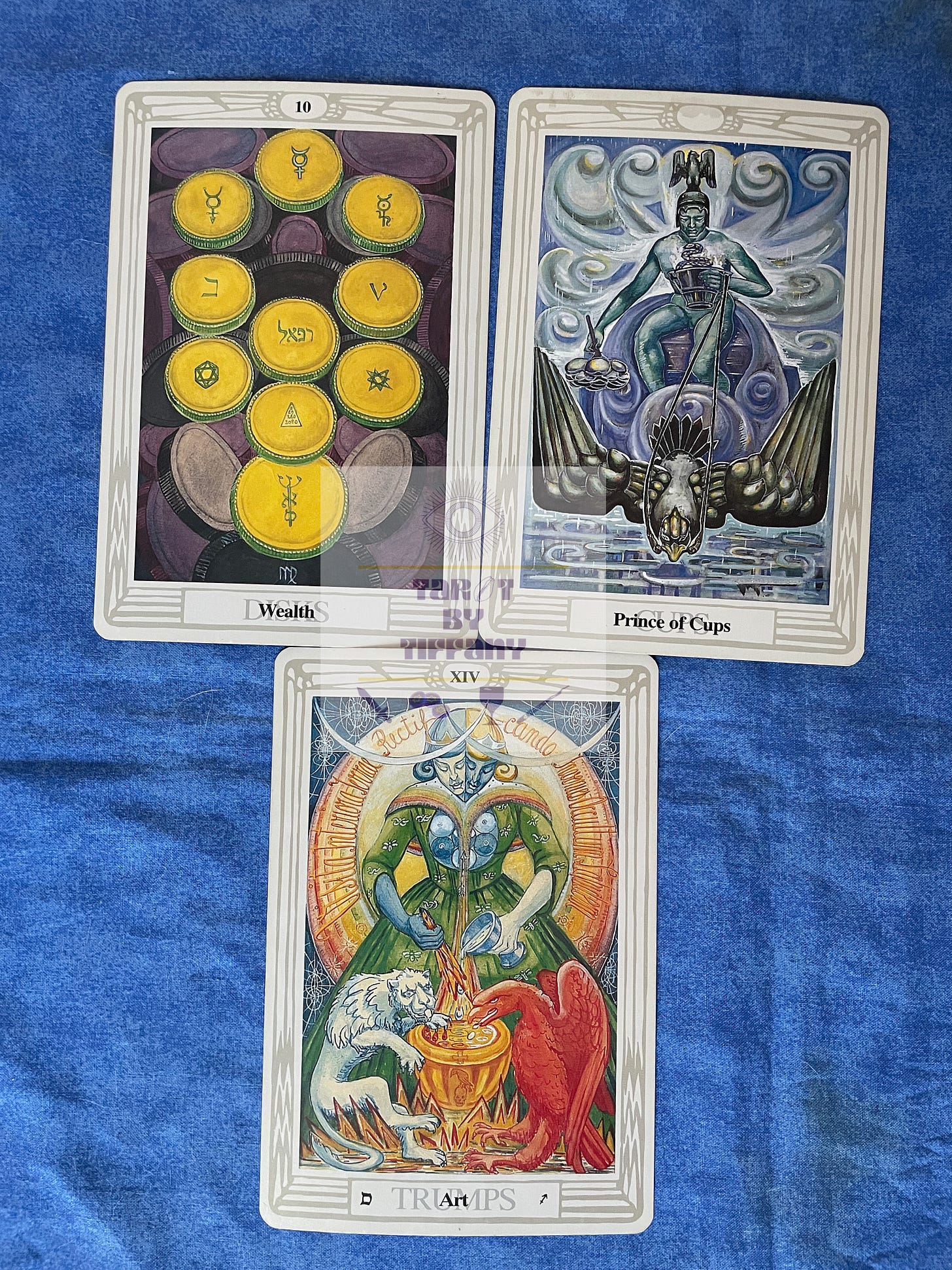 3-card reading with the Thoth Tarot. The cards are laid against a cobalt blue cloth background. Top two cards (L-R): 10 of Disks, Prince of Cups. Bottom card: XIV - Art.