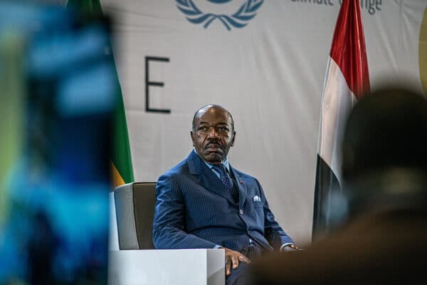 A man wearing a blue pinstripe suit sits in a grey-and-white chair in front of a U.N. banner and several flags.