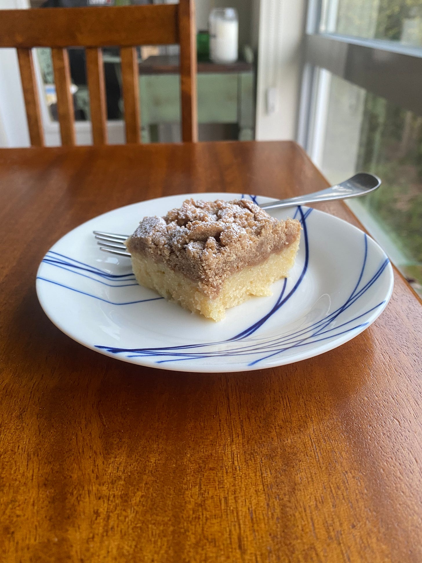 A slice of the crumb cake described above, on a blue and white plate with a fork resting at the back. The cake is equal parts yellow vanilla cake and light brown crumb topping, dusted with powdered sugar.