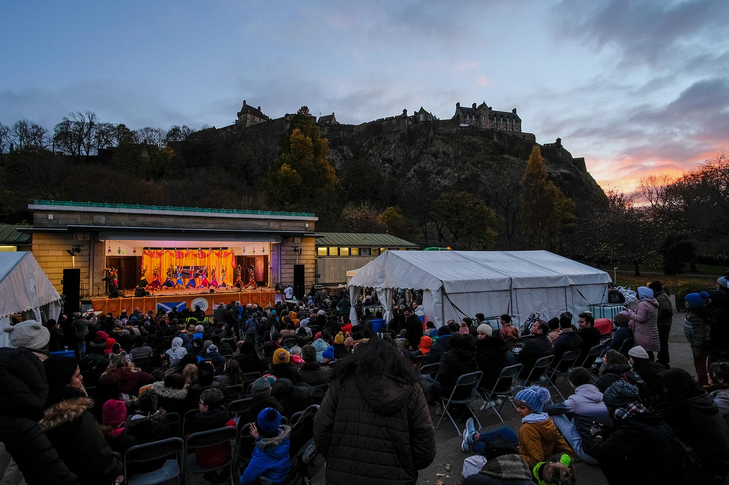 crowd seated in princes st gardens with edinburgh castle in the background. It is dusk and there are Indian dancers on the stage