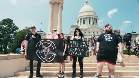 Young people dressed all in black on the steps of a capitol building hold a flag with a pentacle, and a sign that says Religious Liberty for All.