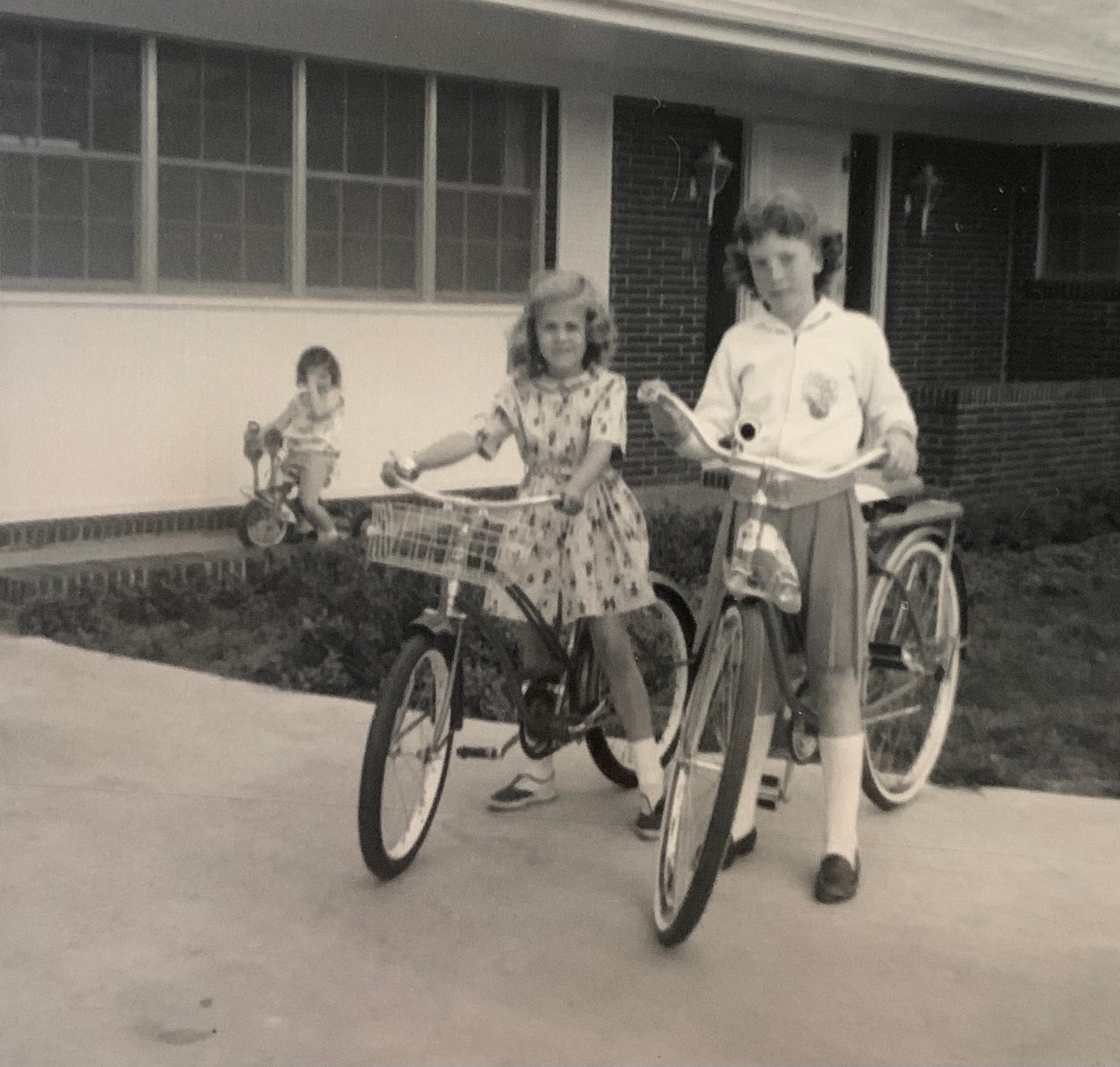 Two girls on bicycles and one on a tricycle in front of a house