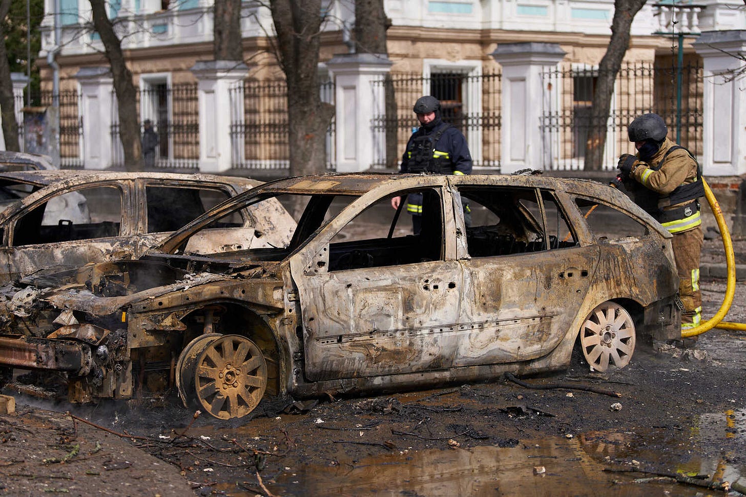 Two emergency workers stand behind burned-out vehicles.