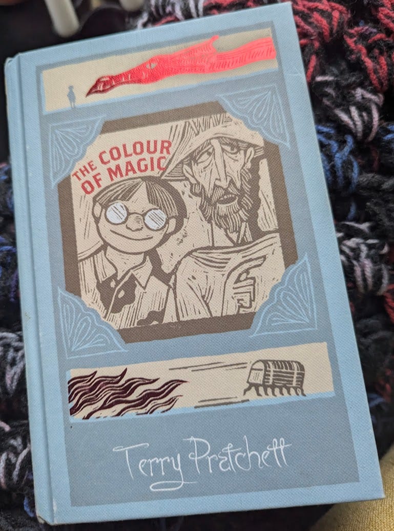 An image of the hardcover copy of The Colour of Magic. It's blue with a wood-cut-like image of Rincewind and Twoflower on the cover.