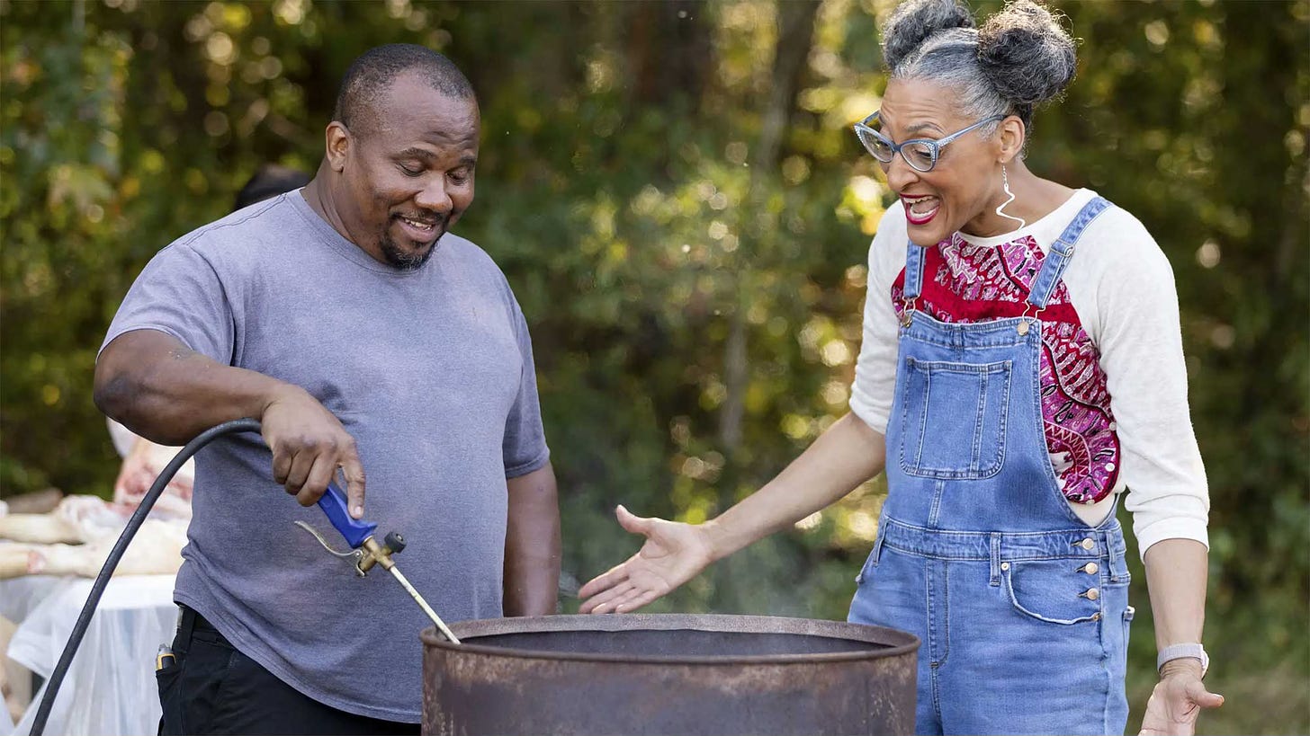 Chef Carla Hall standing over a fire pit with a pit master.