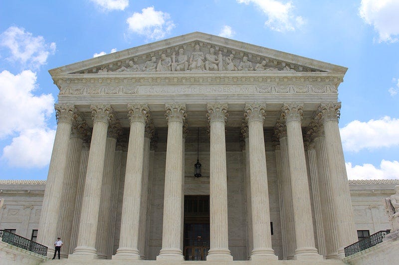 The U.S. Supreme Court building, viewed squarely, under a partly cloudy sky. A security guard stands to one side.