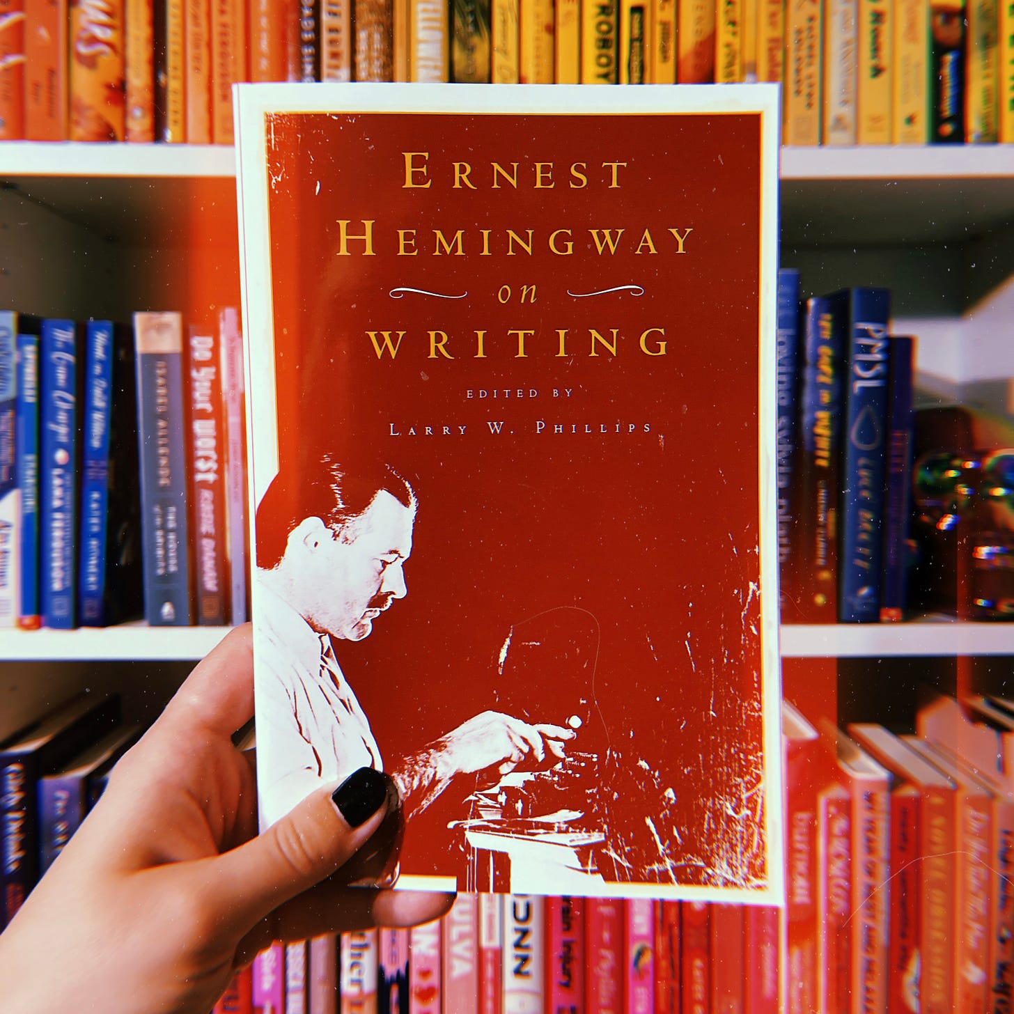 A white woman's hand with black fingernails holds a copy of Ernest Hemingway on Writing, edited by Larry W. Phillips.