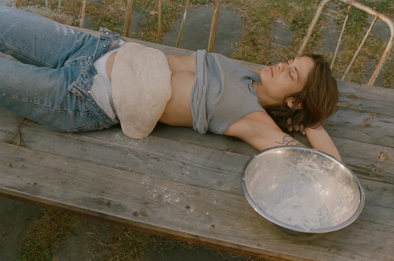 bread artist lexie smith laying down on a table with a sourdough across her stomach