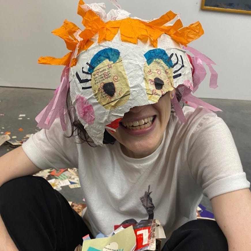 a portrait of a white woman sitting on concrete floor. She wears a white t-shirt and has a huge smile; on her head, she wears the remains of a pinata with cartoon "eyes" and tissue paper "hair"